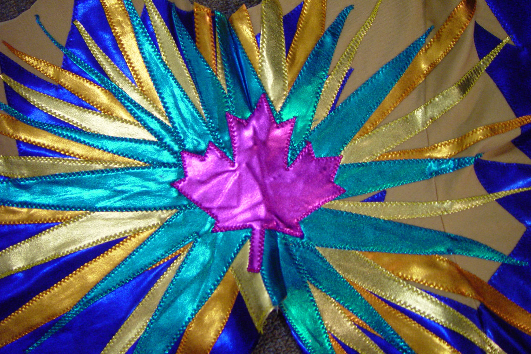 A close up view of intricately appliqued spandex. Shiny foil lycra makes a starburst design, coming out from behind a pink maple leaf.