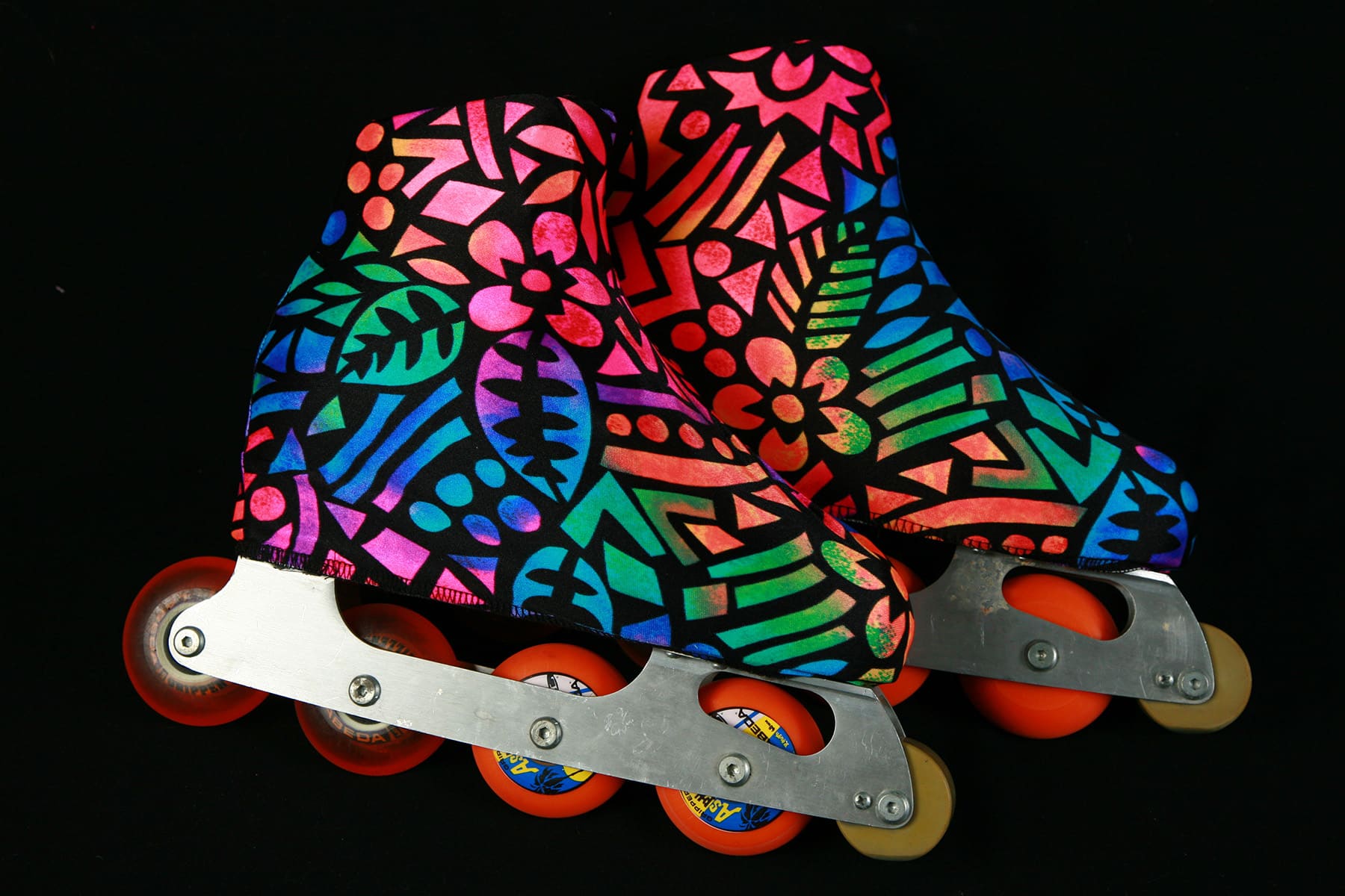 A pair of inline figure skates, in front of a black background. The skates have orange wheels, and are sporting skate covers with a brightly coloured, wild geometric pattern.
