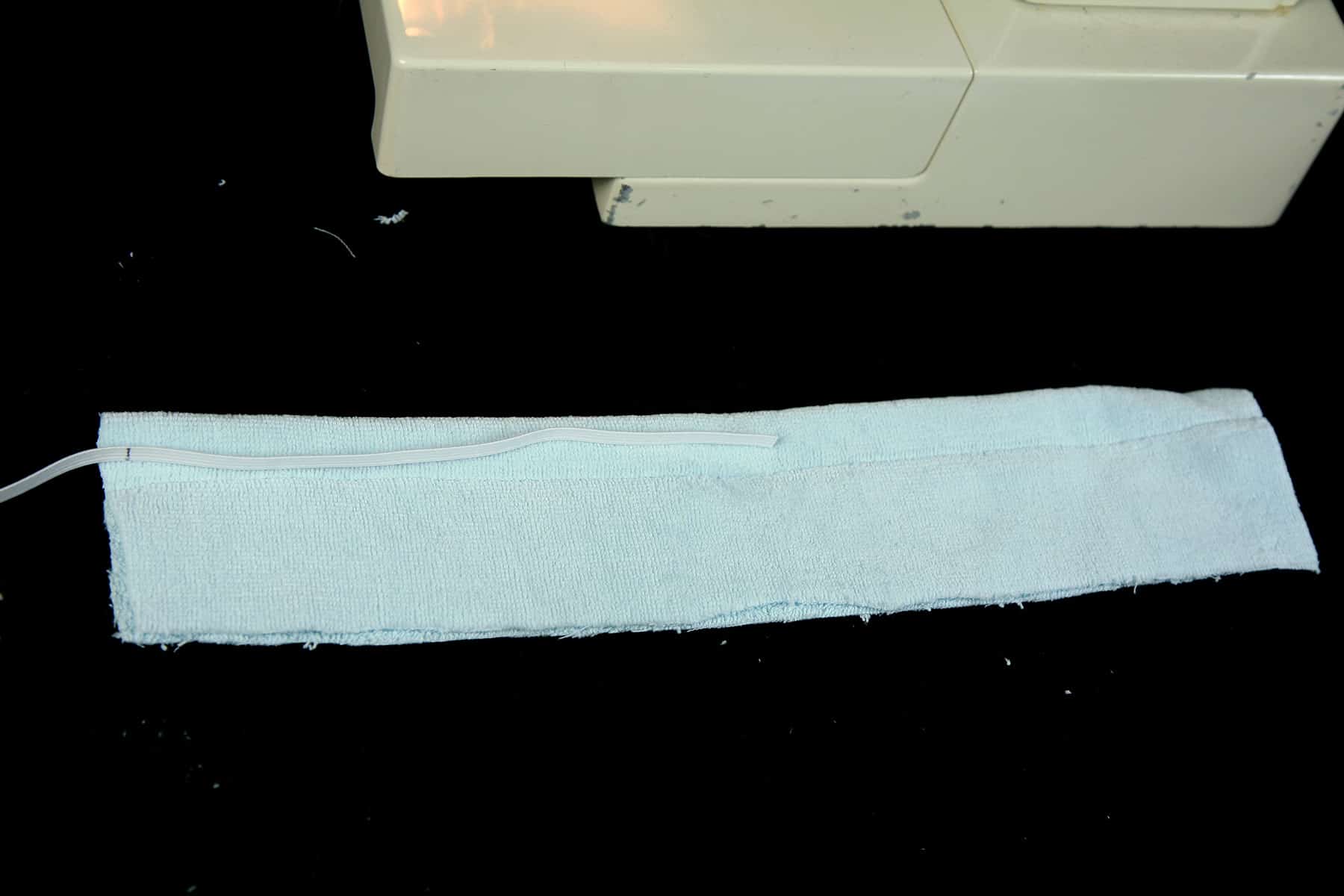 A strip of light blue terrycloth is laying in front of a sewing machine. There is a length of white elastic laying on top of the fabric.