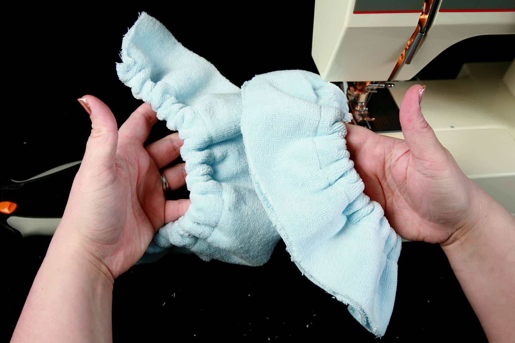 A pair of hands hold up two gathered sections of light blue terrycloth in front of a sewing machine.