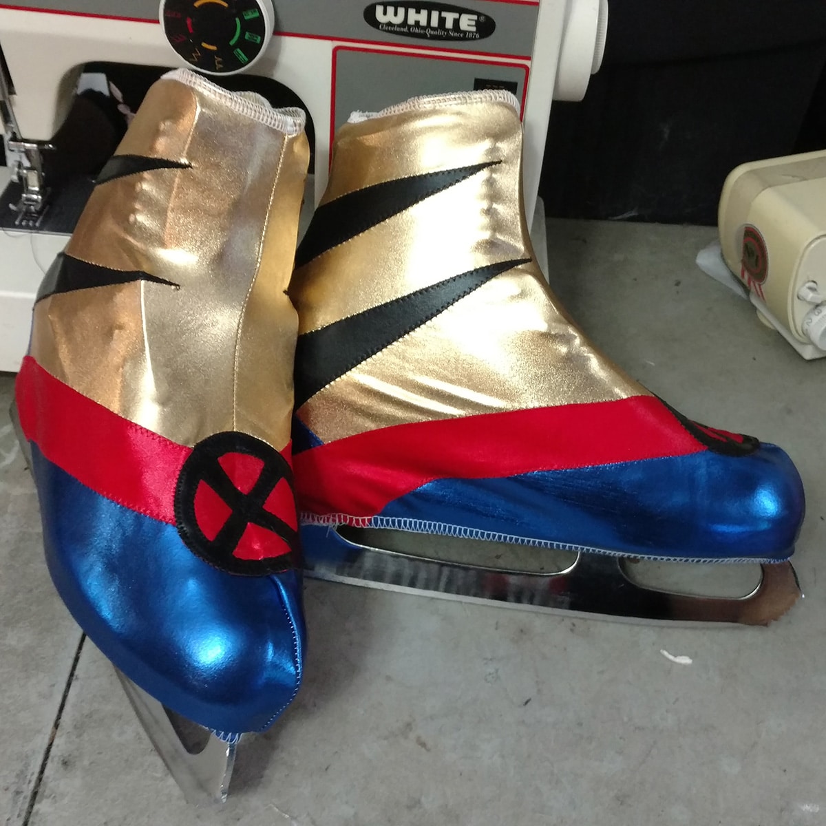 Gold, red, and royal blue skate covers with an X-Men logo and Wolverine slashes are shown on a pair of figure skates.