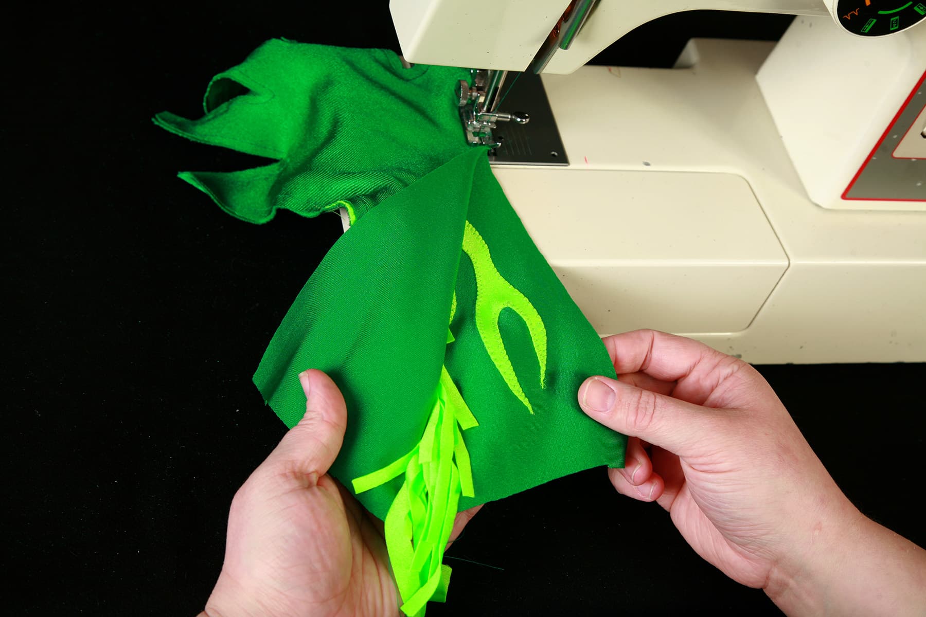 A dark green glove with bright green fringe is being sewn by a sewing machine.