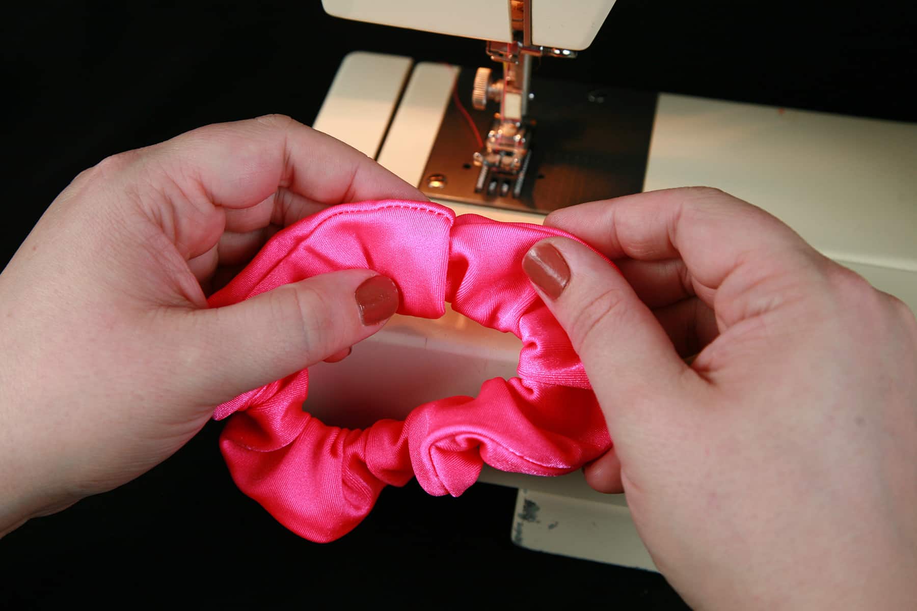 Two hands are shown manipulating a scrunchie in front of a sewing machine.