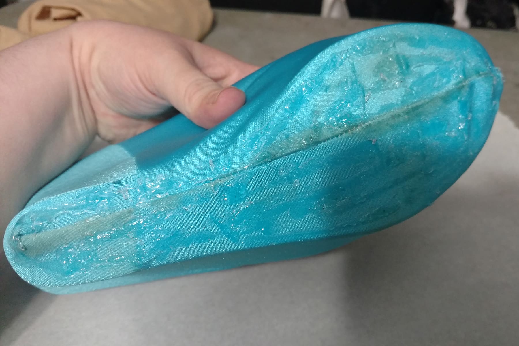 A hand displays the bottom of a blue fabric covered shoe. The bottom is covered in a thick, clear glue.