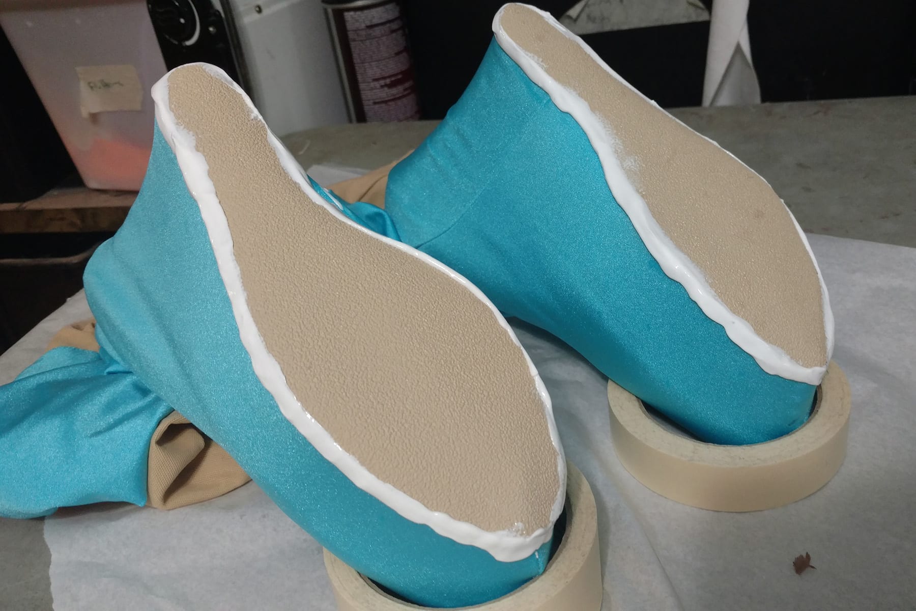 Two shoes covered in blue fabric are shown upside down, the toes balanced on rolls of masking tape. The beige soles are edged in wet caulking, appearing white.