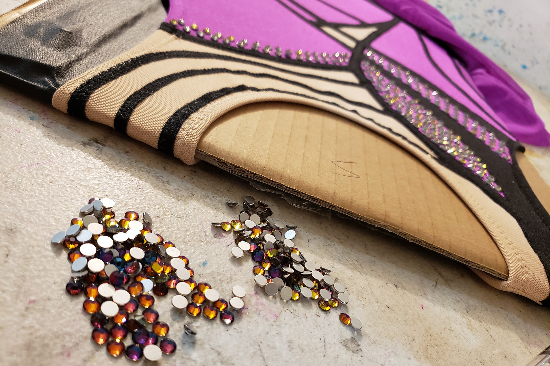 A close up view of the purple and black skating dress on a cardboard stretcher, with a pile of purple-blue rhinestones in front of it.