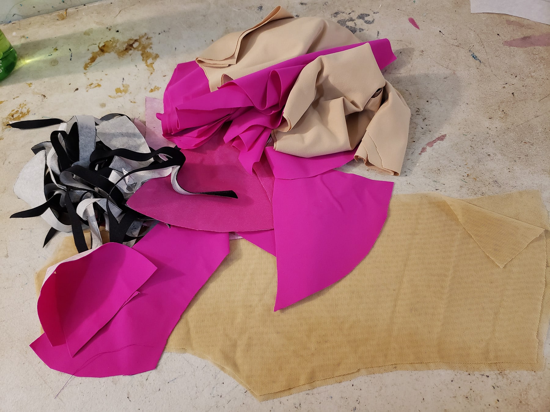 A small pile of cut fabric pieces on a work table. There are pieces of hot pink spandex, hot pink mesh, beige mesh, beige lining, and black spandex.