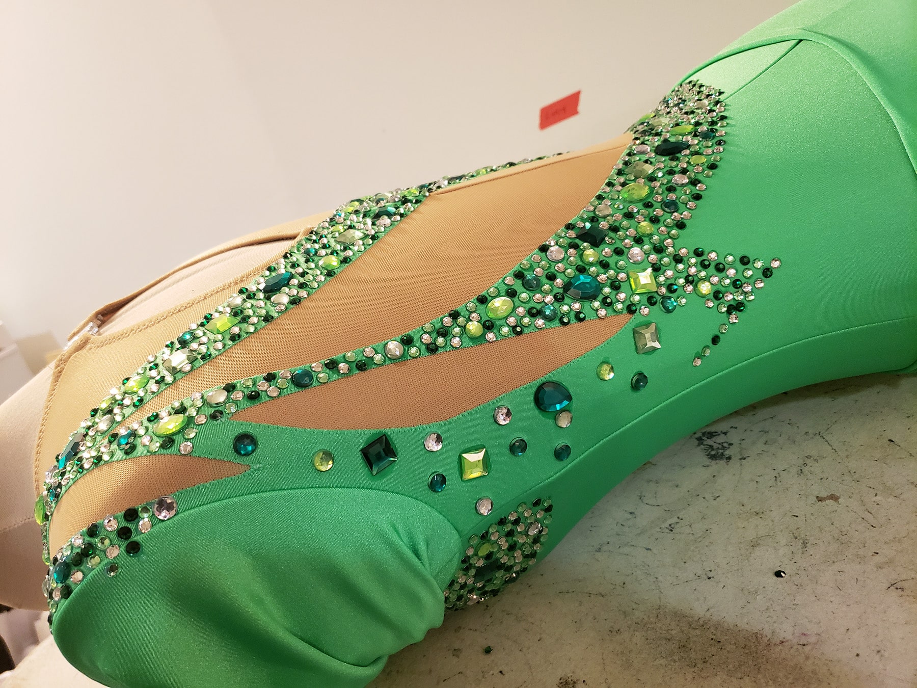 A green skating dress on a dress form, with large rhinestones glued on. The rhinestones are clear and various shades of green, in a variety of shapes and sizes.