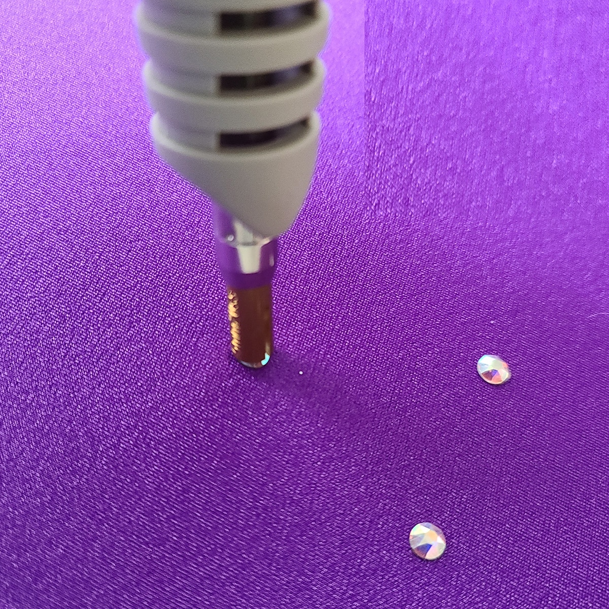 A hotfix iron is applying a crystal to a piece of purple spandex.