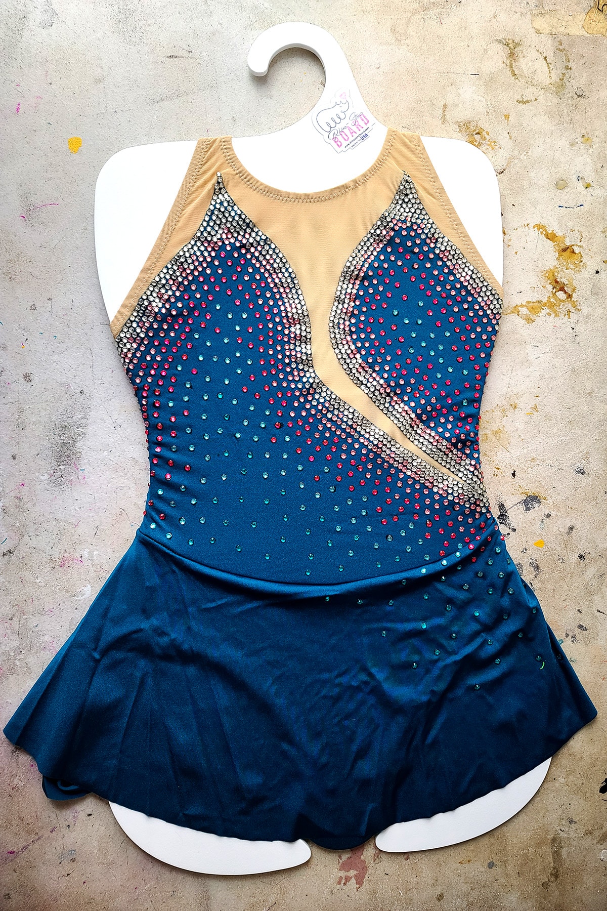 A dark teal skating dress with multicoloured crystals is stretched on a Rhino-Stone Board