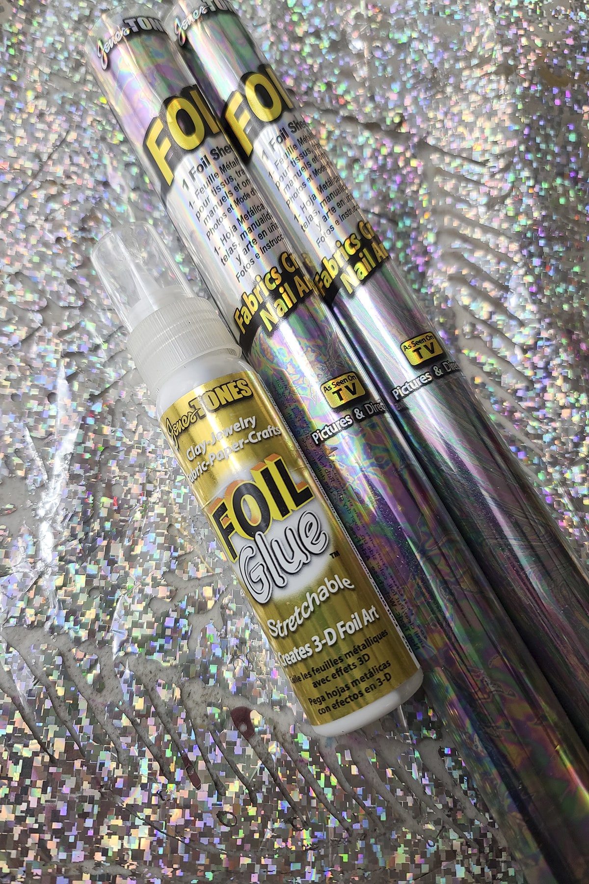 Two rolls of holographic foil paper and a bottle of foil glue, resting on a sheet of holographic foil.