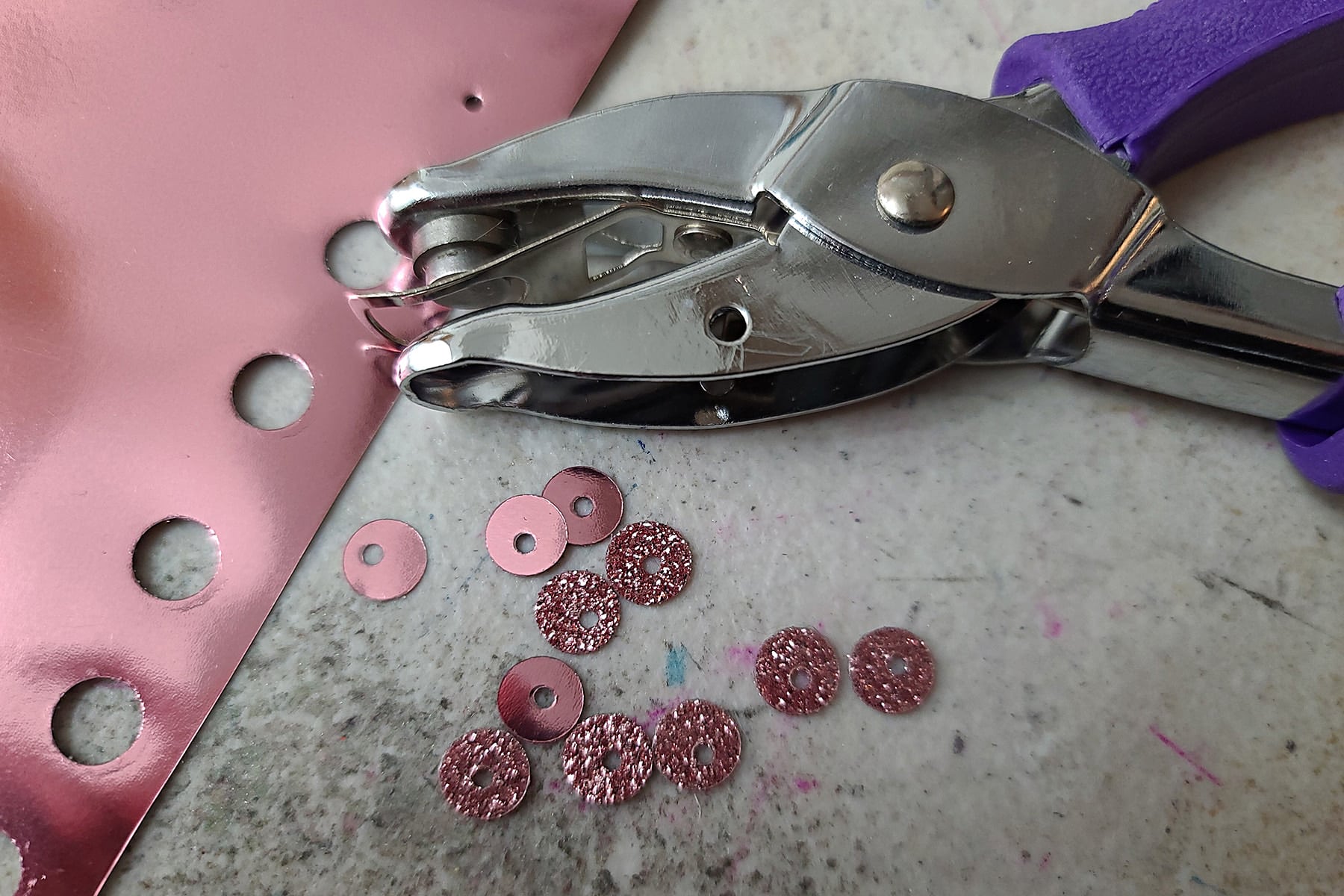 A hole punch is being used to make sequins. There is a small pile of pink sequins on the surface below the hole punch.