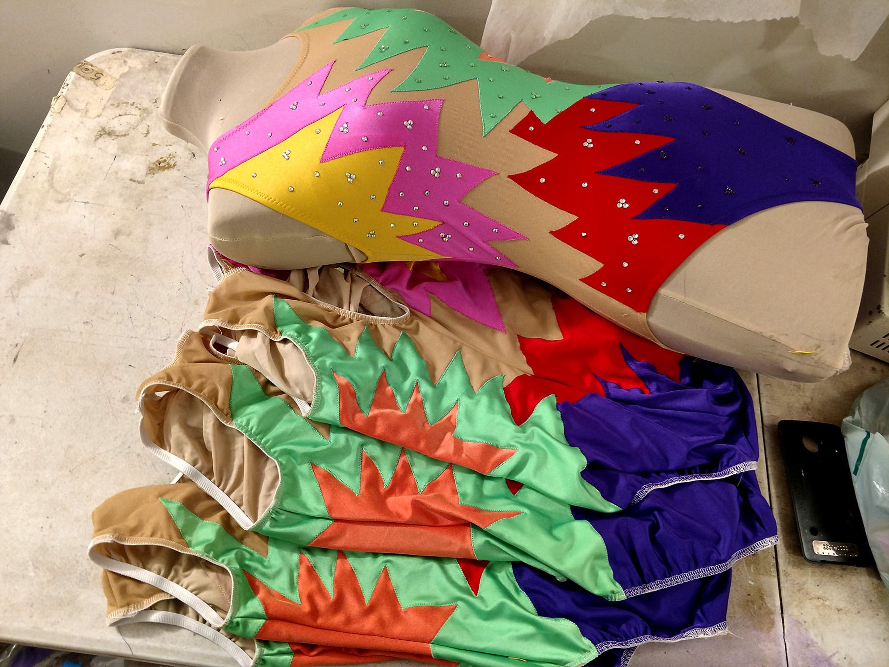 A pile of jazz team synchro suits is shown. They are mostly done, the elastic is yet to be flipped and stitched down. Beside the pile is a dress form with one completed suit on it.