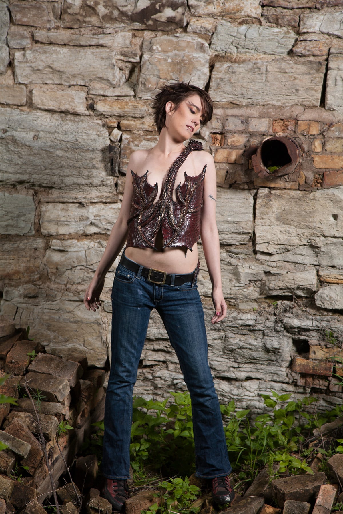 A beautiful young woman with short hair is standing in front of a rock retaining wall, wearing a dark burgundy latex, 3D sculpted dragon top. It wraps around her torso, and looks like Smaug.