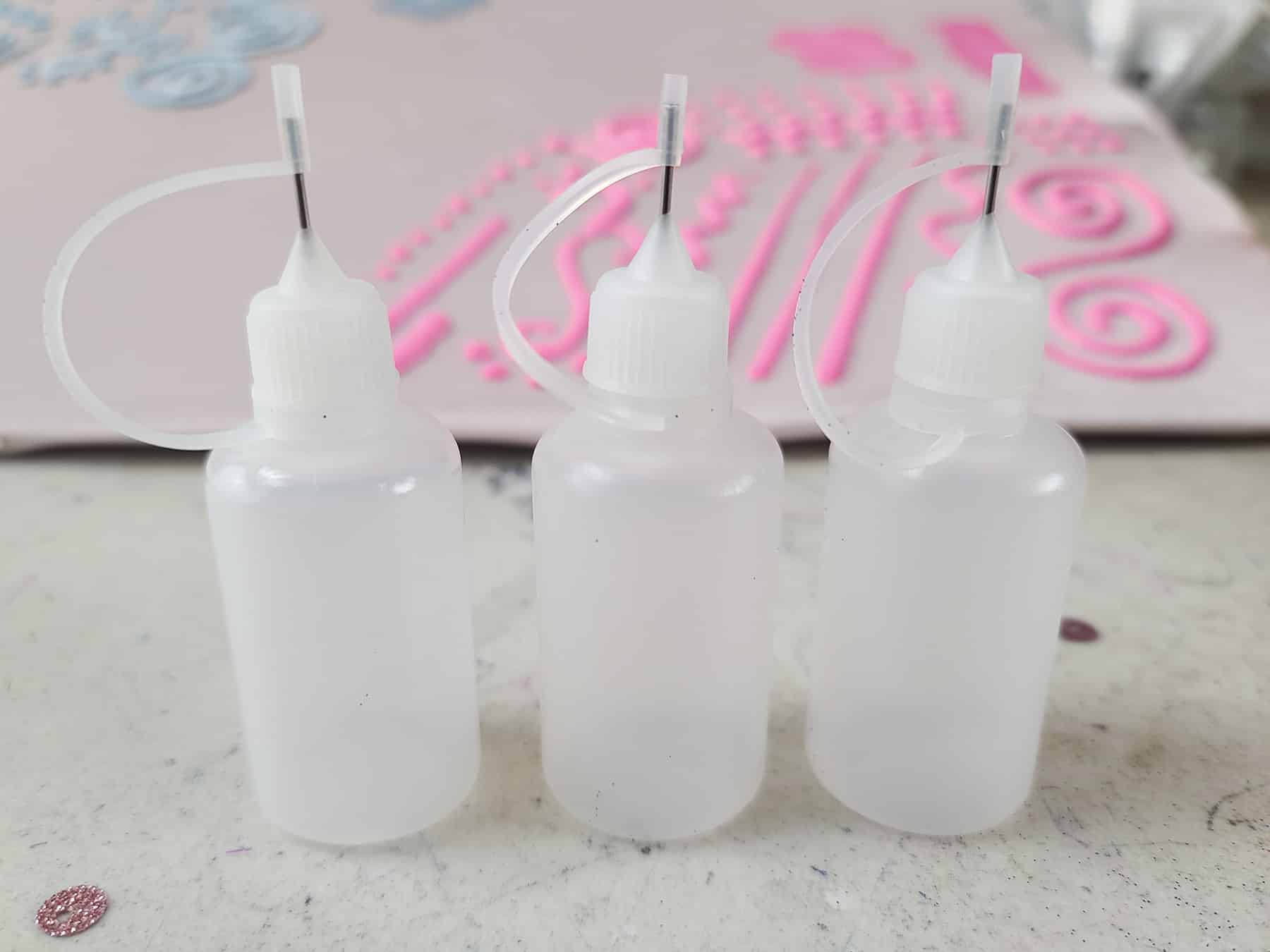 3 small translucent white empty fabric paint bottles in front of a piece of light pink spandex with neon pink stretch paint designs on it.