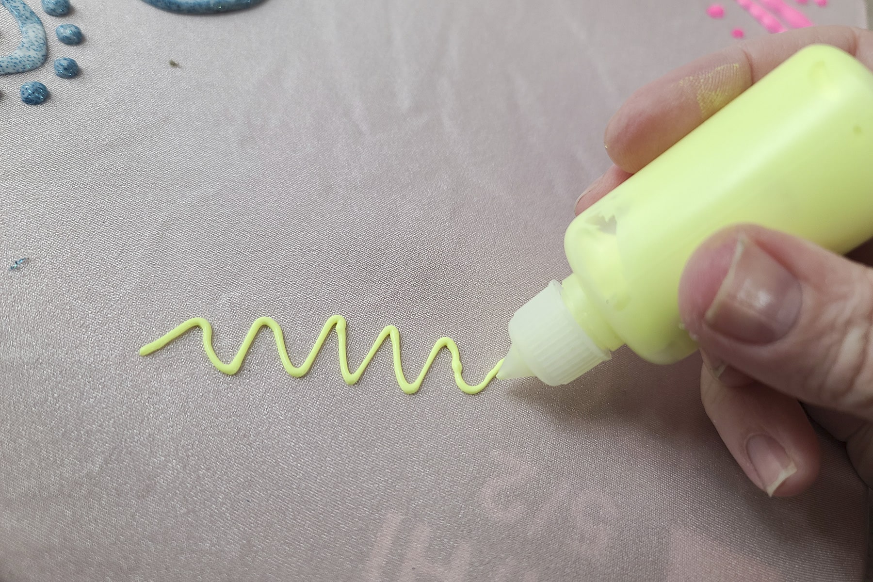 A small fabric paint bottle with neon yellow stretch paint is piping a squiggle onto light pink spandex.