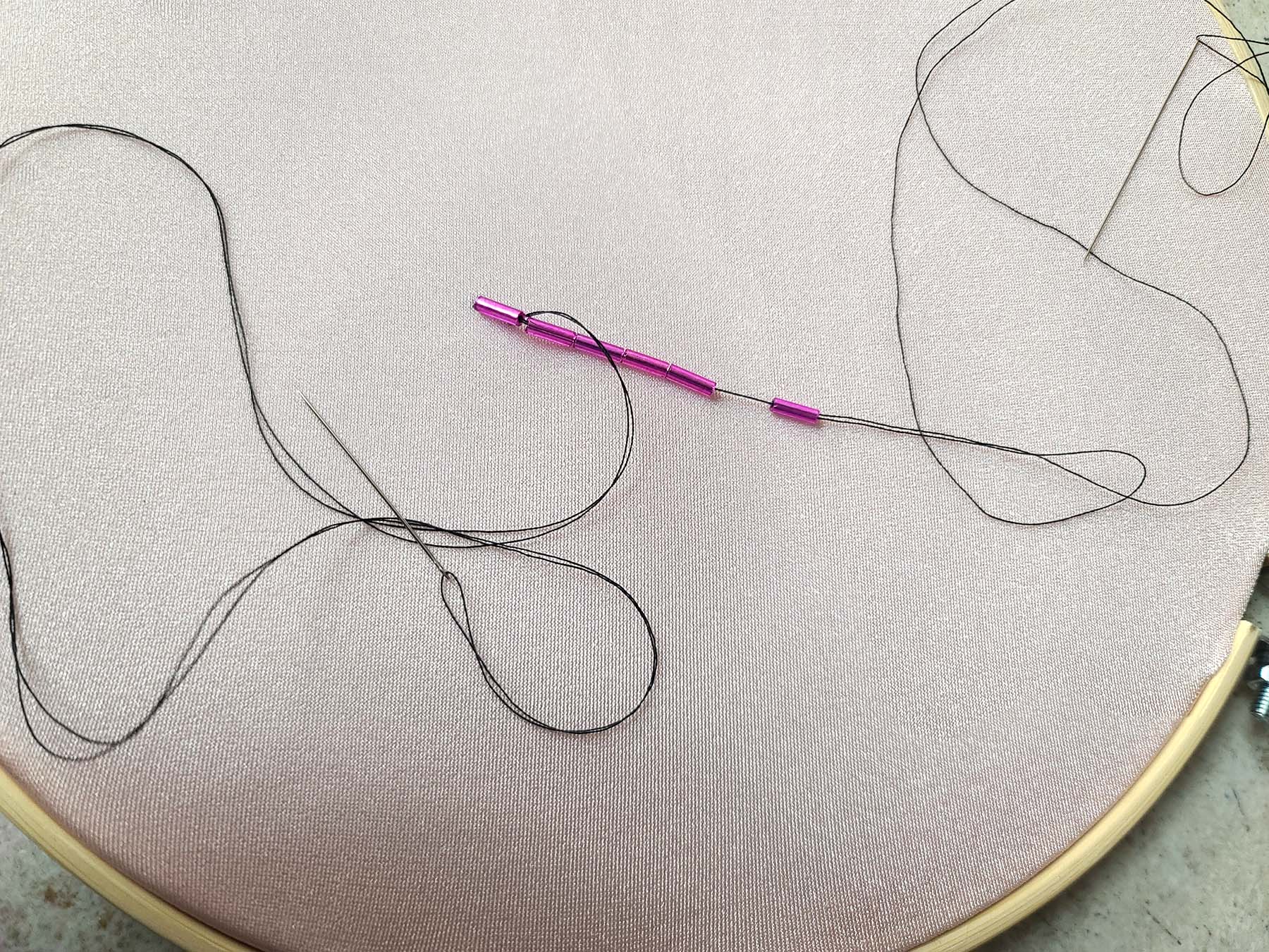 Dark pink bugle beads are threaded onto a black thread, over a light pink spandex. A second needle with black thread is pictured.