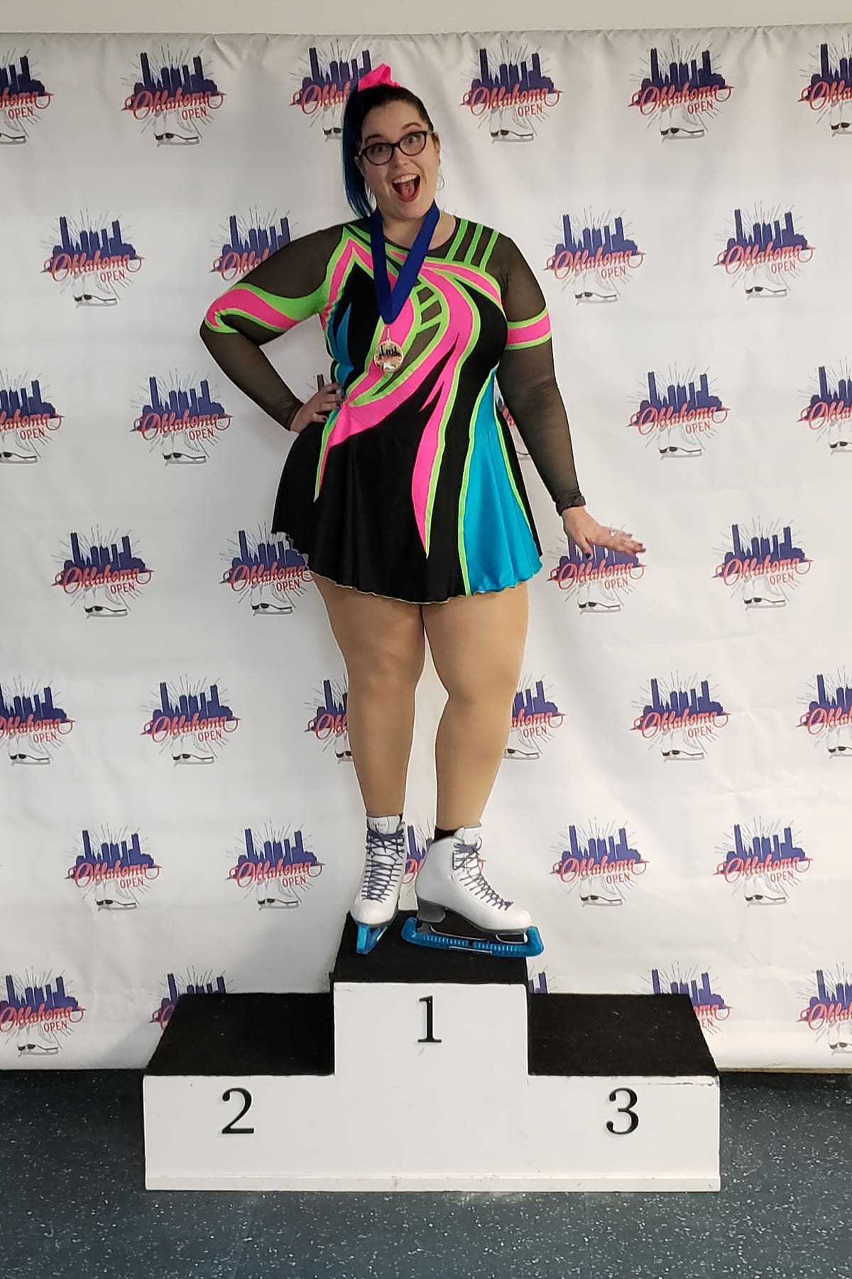 A gorgeous dark haired figure skater in a black, hot pink, lime green, and sky blue 80s themed figure skating dress. She stands alone on the top of a podium, smiling brightly.