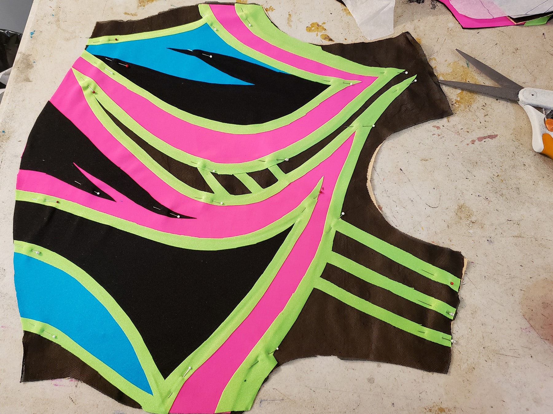 The sewn front bodice of an 80s style figure skating dress. The base is black spandex and mesh, with designs in hot pink, lime green, and sky blue on top.