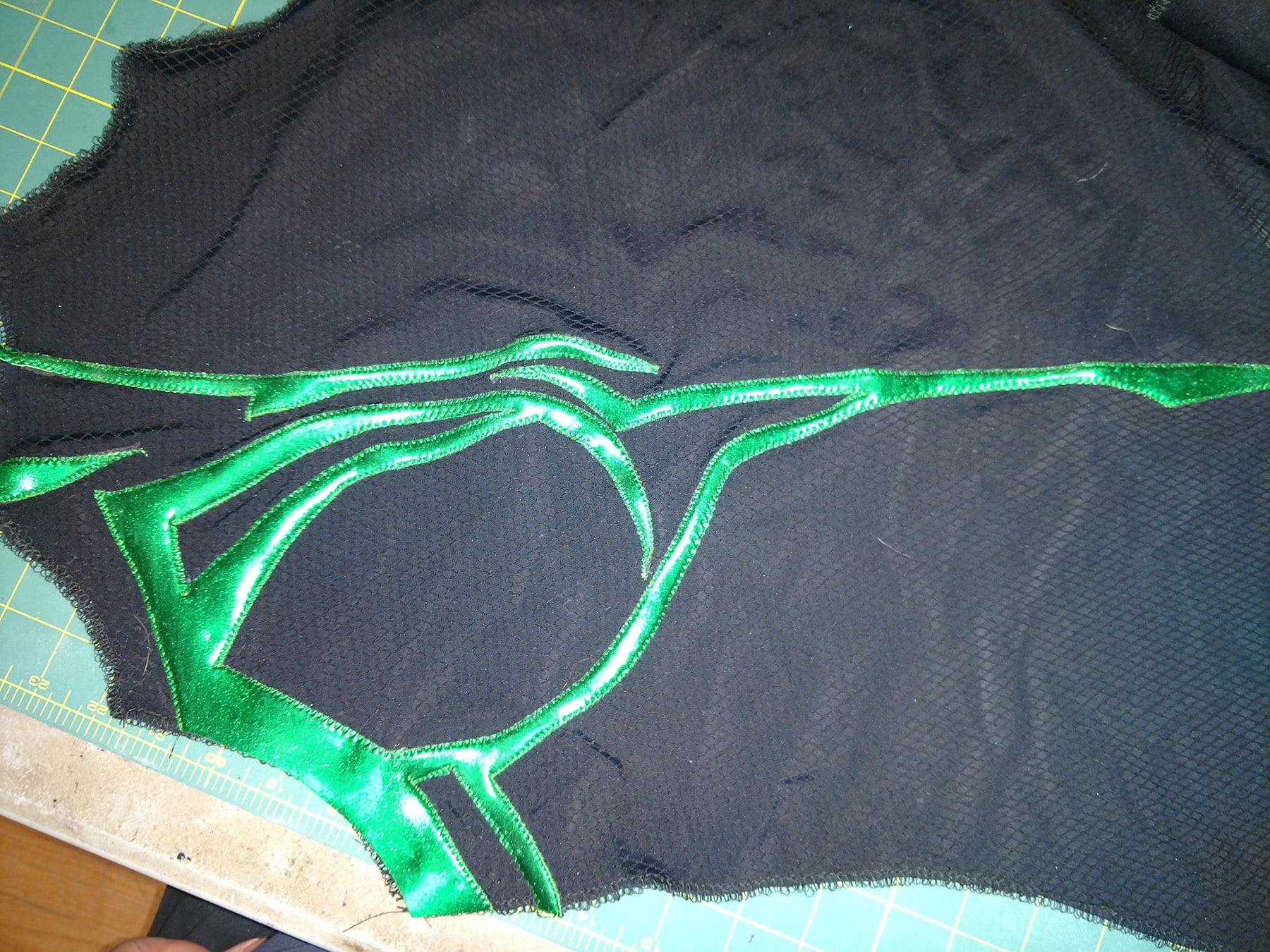 An intricate cutting of shiny green fabric is stitched in place on a textured bodice piece of black spandex.