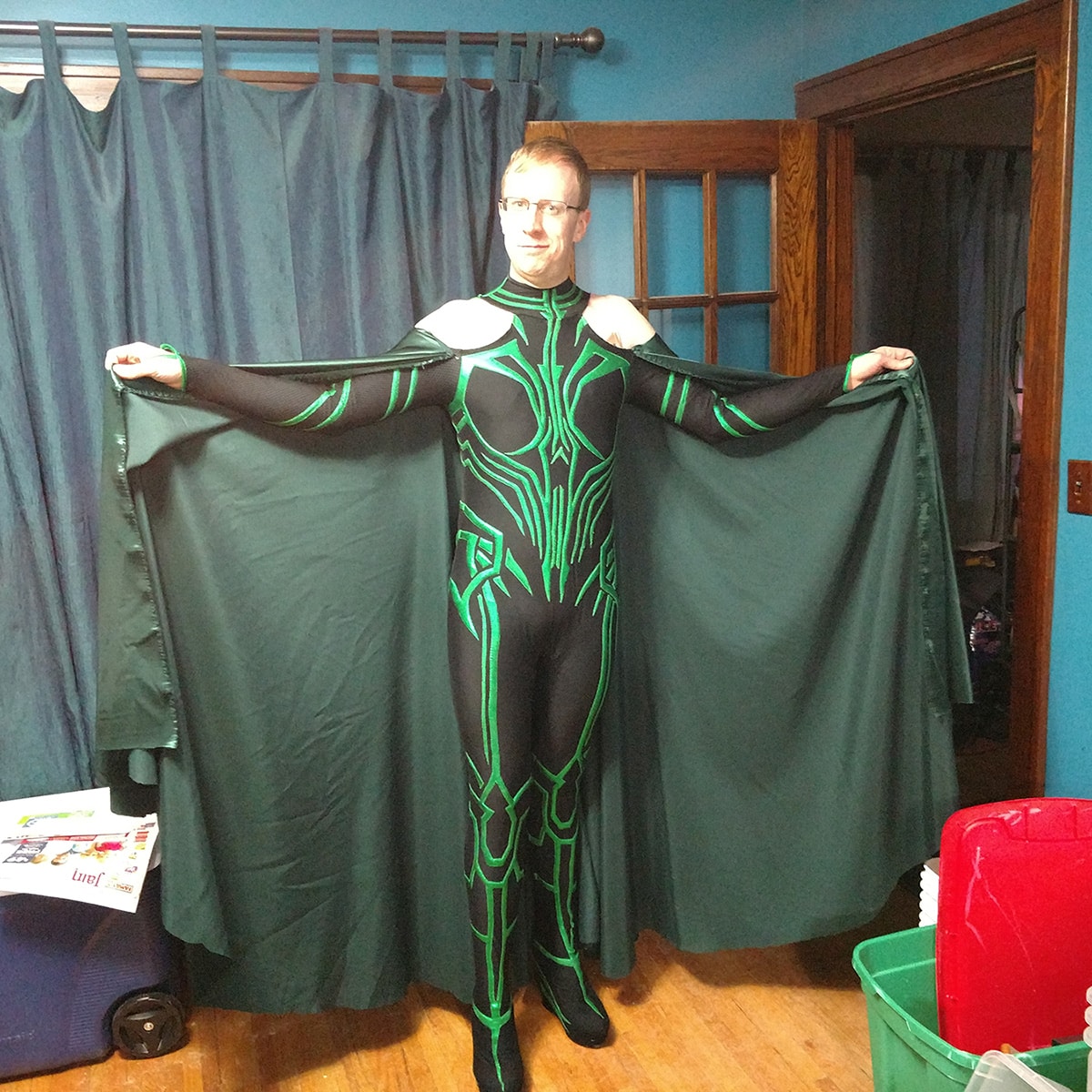 A front view of a blonde man in glasses, wearing an MCU Hela cosplay.  His arms are extended out to the side, holding up the cape ends to show the fullness.