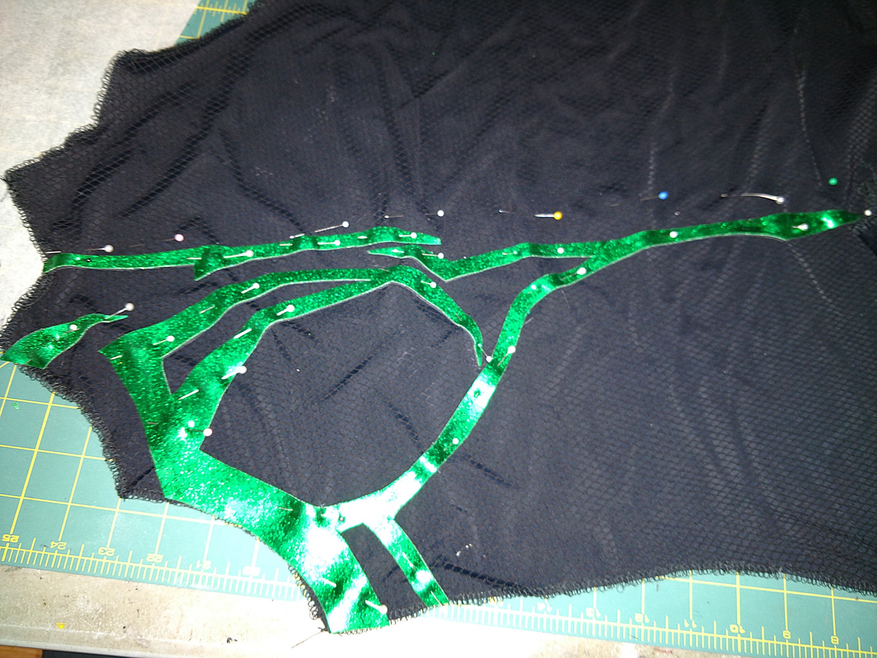 An intricate cutting of shiny green fabric is pinned in place on a textured bodice piece of black spandex.
