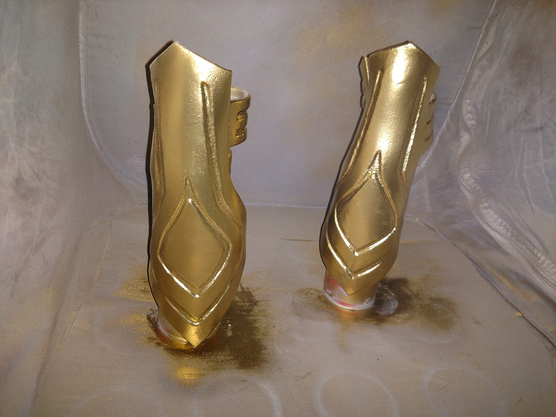 A pair of shiny gold cosplay Hela bracers are shown against a grey backdrop that has gold spray paint on it.