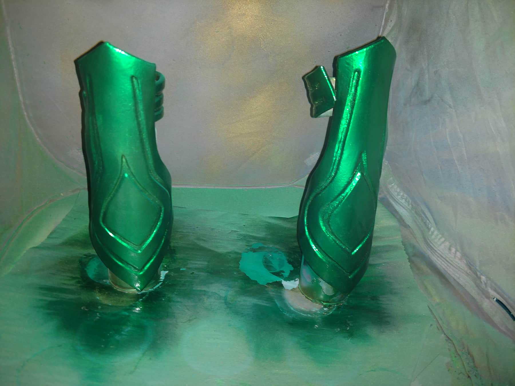 Shiny metallic green Hela bracers are shown against a grey backdrop that is covered in green and gold spray paint.