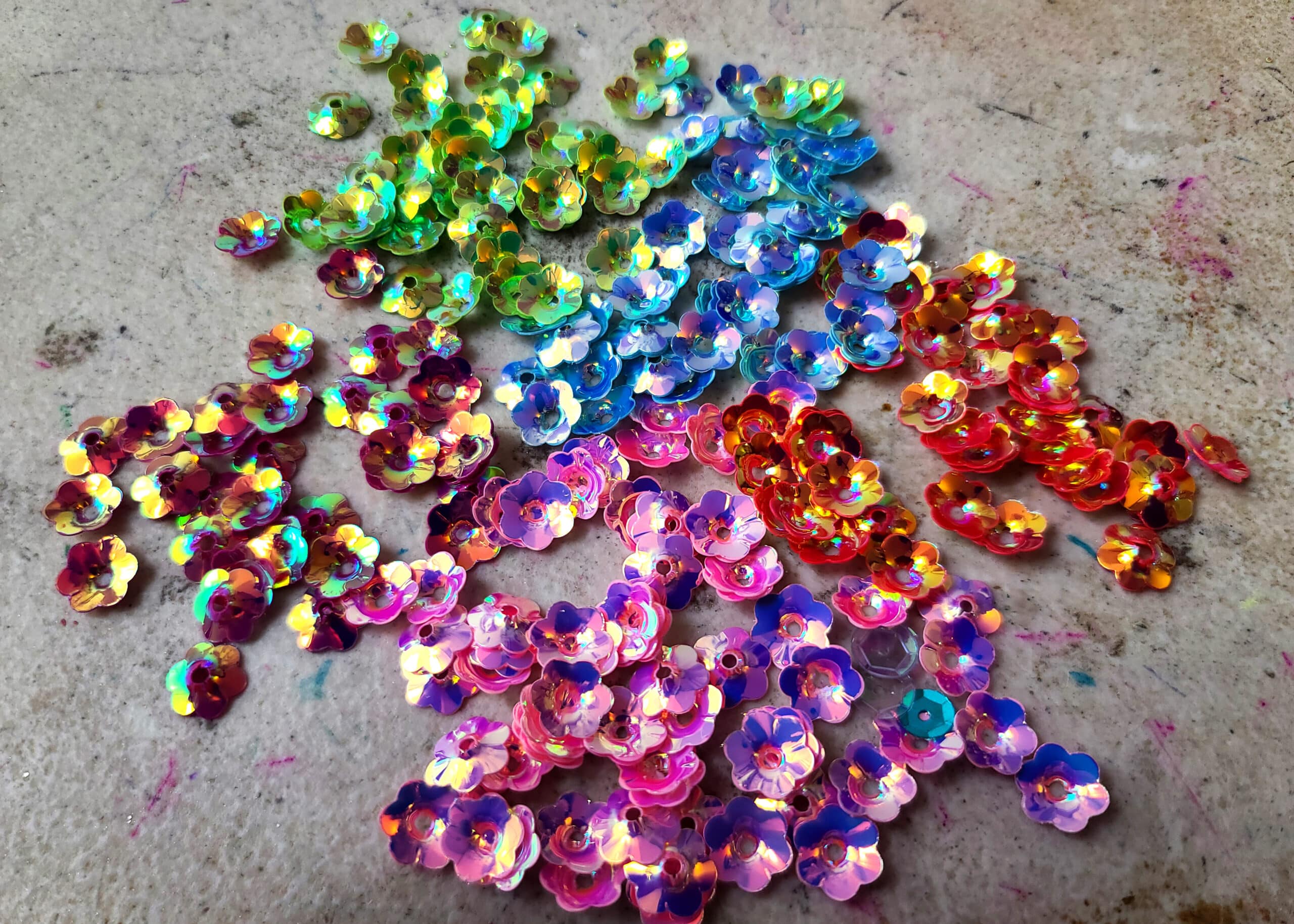 Several piles of iridescent, flower-shaped sequins in pink, orange, blue, green, and purple.