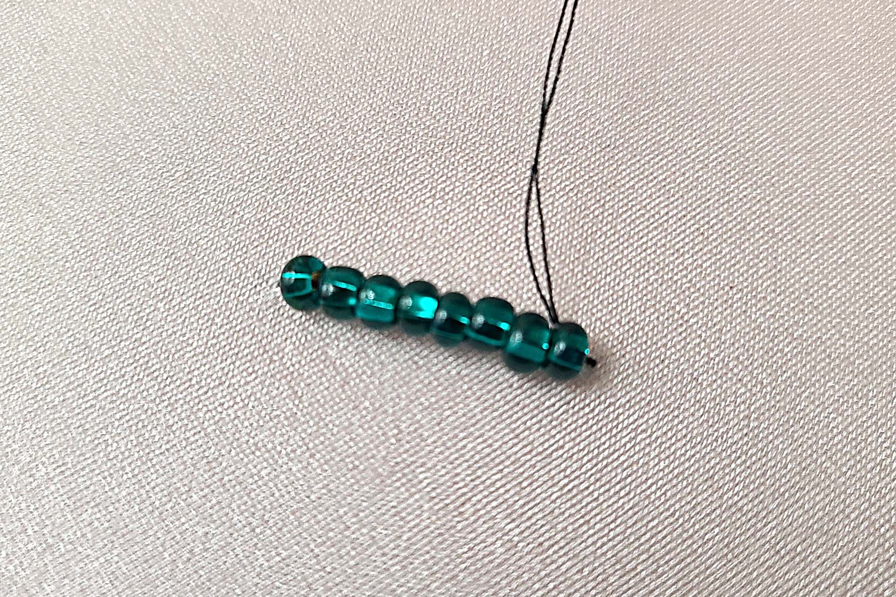 Black thread is being used to sew a short line of dark green rocaille beads to a stretched piece of light pink spandex.