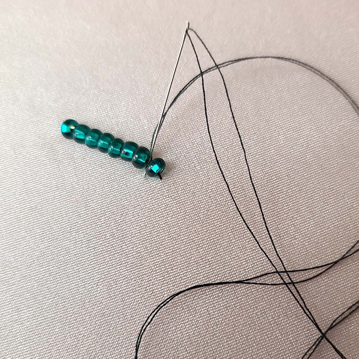 Black thread is being used to sew a short line of dark green rocaille beads to a stretched piece of light pink spandex.
