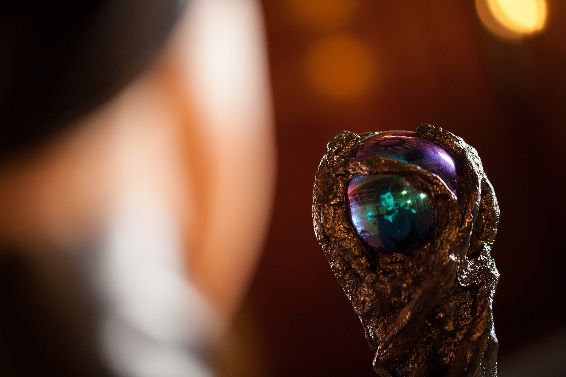 A photo taken over the shoulder of a Maleficent cosplayer. She is looking at the top of her staff, and her reflection is visible in the gazing ball.