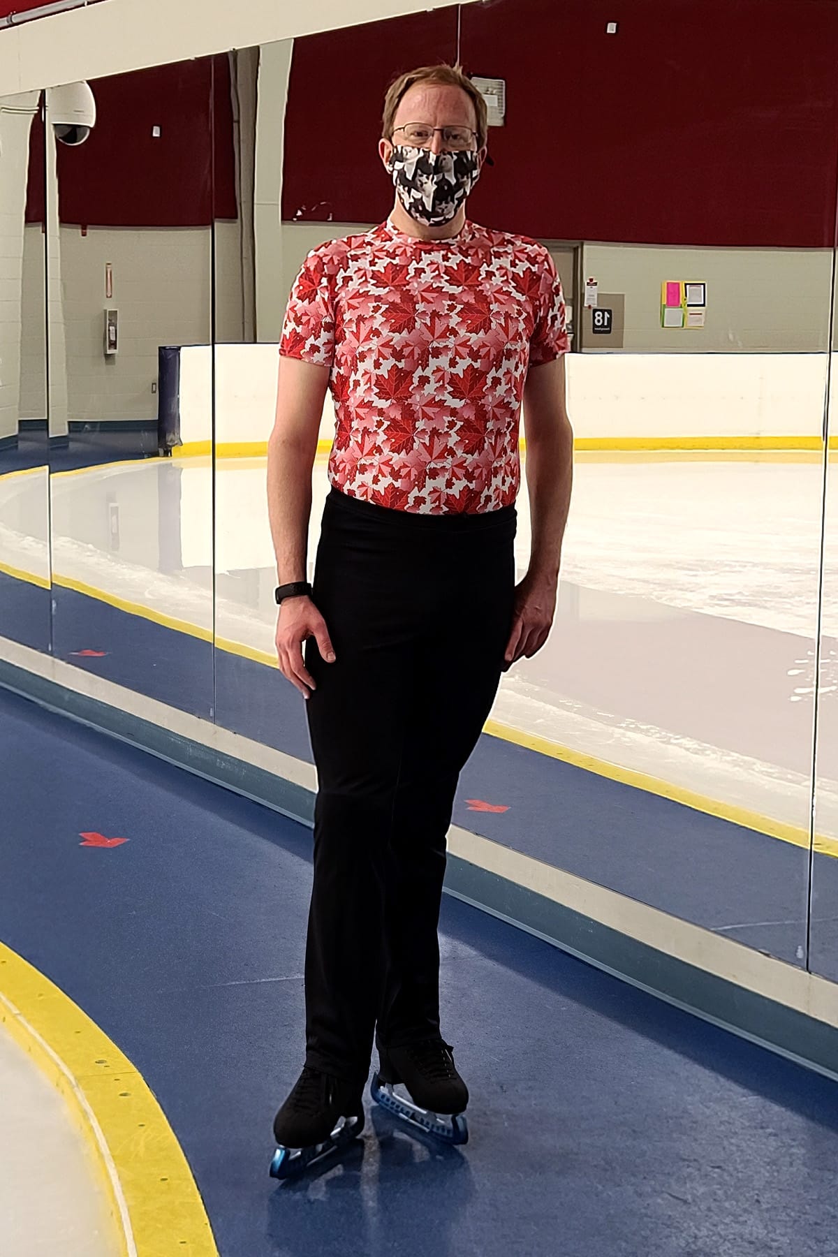 A middle aged man standing at the side of a skating rink, wearing a red and white body shirt with black skating pants.