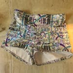 A pair of holographic booty shorts on a sewing table. They are white with turquoise, purple, and black gymnastics words all over them.