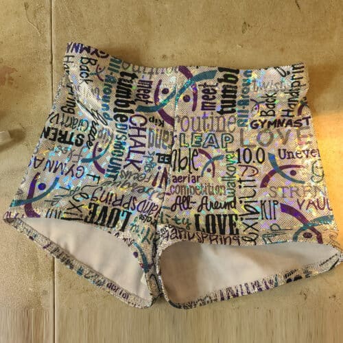 A pair of holographic booty shorts on a sewing table. They are white with turquoise, purple, and black gymnastics words all over them.
