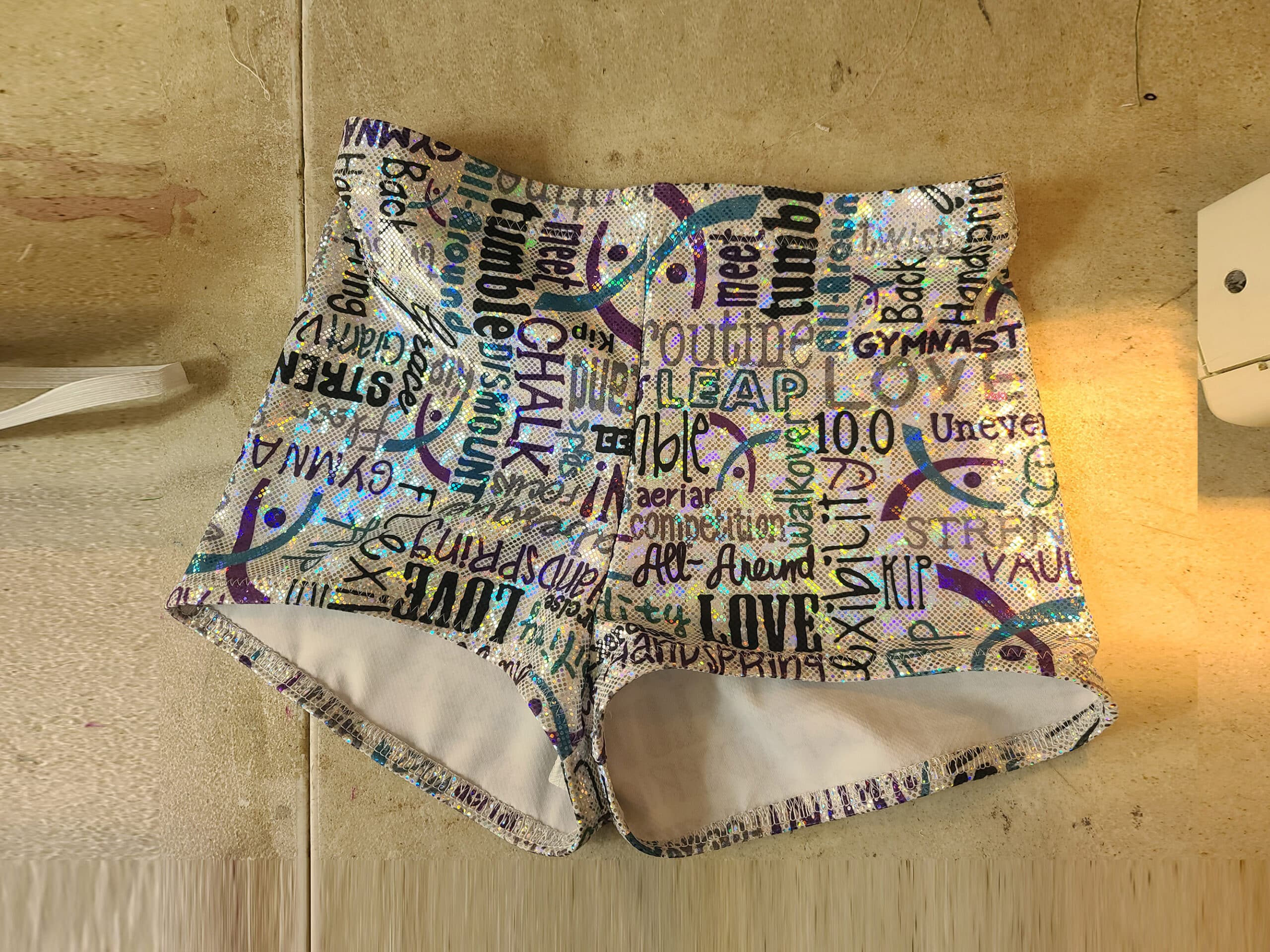 A pair of holographic booty shorts on a sewing table. They are white with turquoise, purple, and black gymnastics words all over them.
